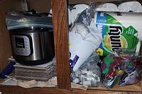 Shelf with Instant Pot Duo 60 V3, stacked cardboard pieces, Bounty paper towels, cookie cutter molds, air freshener beads in plastic bags.