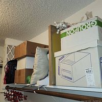 Boxes stacked on shelf including a white box labeled Groupon containing tissue paper, and other cardboard boxes with unknown contents beside a light blue decorative pillow.