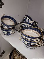 Stack of six double‐handled bowls with blue willow scene and gold lattice band, plus additional cups in background