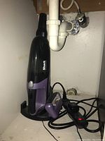 Shark cordless vacuum with charging base and power cord, black with purple accents, placed under a sink.