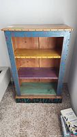 Front view of the wooden bookshelf showing three shelves painted yellow, purple, and teal, with blue and multicolor floral painted exterior and scalloped edge trim.