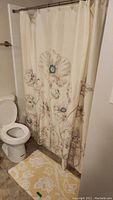 Photo of cream-colored shower curtain with large floral embroidery hanging beside toilet and matching yellow floral bath rug on floor.