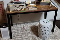 Wood and metal side table with wood-finish top and black metal legs shown with some items on top and beneath on carpet flooring.