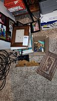 Wooden picture frames stacked along with wooden character figurine, costume jewelry in a small cardboard box, black metal jewelry tree stand visible on carpet.