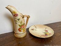 Photo showing the antique Royal Worcester porcelain ewer jug with floral decoration and accompanying matching plate on a wooden surface.