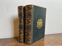 Two volumes standing upright, one showing spine and one showing front board with gilt crest.