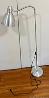 Full view of a silver aluminum modernist floor lamp with a slender vertical stand, round base, and black power cord with floor switch.