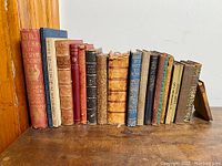 Photo showing full collection of 18 antique books on a wooden surface, varying in size and color from reds, blues, and browns, with worn bindings and gilt lettering.