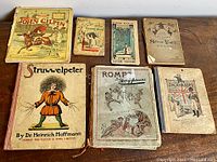 Seven antique children's books displayed on a wooden surface, showing wear and some loose pages.