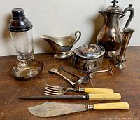 Overall view of antique silver plated items including teapot, gravy boat, vase, cocktail shaker, sugar bowl, decorative dish, and flatware with ivory handles.