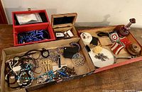 Wide shot showing various costume jewelry pieces arranged in boxes and a carved wooden box with a cranberry glass bottle with stopper nearby.