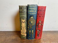 Front view of the spines of three G.A. Henty illustrated antique books showing distinct illustrations and titles.