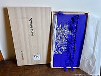 Wooden box with Japanese writing on top holding the folded blue silk tapestry with silver samurai illustration partially visible, original wrapping inside box