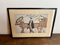 Framed woodblock print titled 'Le Depart' by Harunobu showing three women in traditional Japanese dress. View of entire framed piece leaning on a wooden surface.