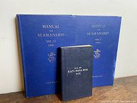 Three vintage sailor's manuals arranged against a wall: two large 1951 Manuals of Seamanship Vol I and III in blue, and one smaller 1935 Boat's Signal Book in black.