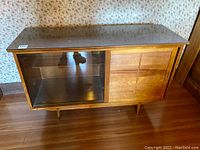 Front view of the vintage mid-century modern wood cabinet showing sliding glass door on left and wood door on right, standing on tapered legs.