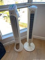 Two white tower fans standing side by side near a window, both approximately 39 inches tall, one with control buttons on top and the other with a vented grill.