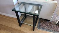Front angle photo of rectangular glass side table showing clear glass top, lower glass shelf, and black metal frame.