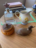 Overview of the glass decanter with star lid, ceramic milk jug with floral pattern, wooden trinket box with carved leaf and knob design on lid, and carved wooden duck figurine.