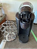 Front view of black Keurig coffee maker with pod compartment open, next to silver metal coffee pod holder on kitchen counter