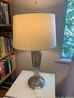 Full view of the lamp showing metallic base and white shade on a white table near a window.