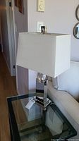 Front and side angle views of a silver metal base table lamp with rectangular white fabric shade and clear accent section near the top of the base, placed on a glass topped table next to a white couch.