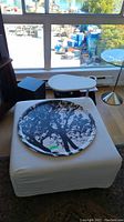 Ikea ottoman with white fitted cover, decorative round black and white tree silhouette tray and small white side table attachment next to large window.