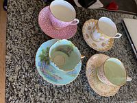 Four different tea cup and saucer pairs arranged on granite countertop, showing various floral and scenic patterns with gold trim