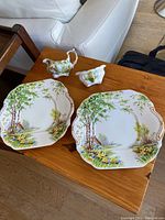 View of two tea plates, creamer, and sugar bowl on wooden table showing nature scene and floral patterns.