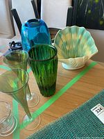 Photo shows blue glass vase, green ribbed glass vase, two green opalescent champagne flutes, and shell-shaped ceramic vase on a wooden table.