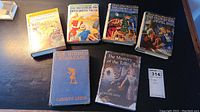 Six vintage Nancy Drew books with both dust jackets and cloth bindings, titles clearly visible, laid out on a wooden table.