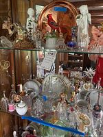 Glass and ceramic bells, plates, and figurines displayed on glass shelves with wooden background. Includes copper plate, various angel and bird figurines.