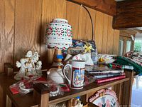 View of lamp with wooden base, decorative holiday shade, white ceramic mug with Christmas design, angel figurines, packaged Christmas linens, and books on shelf