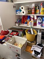 Photo of white kitchen cabinet with cleaning supplies including paper towels, cleaning sprays, laundry detergent, yellow bucket, electric toothbrush and some household items.