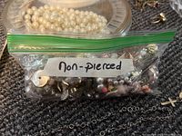 Clear plastic bag labeled 'non-pierced' containing various clip-on earrings, including metallic and pearl elements, set on a gray carpet with some chains visible underneath.