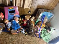 Wide angle image showing entire lot of assorted plush toys, baby dolls, paper doll, and a Cookie Monster face cutout stand on carpeted floor against paneled wood wall.