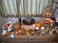 Wide view showing fall holiday themed figures including pumpkins, pilgrim figurines, ceramic pitchers, a horn of plenty, and a wooden cheese board on a dresser.