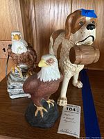 Three figurines displayed on wooden shelf including two eagle figurines and one St Bernard dog figurine with barrel