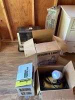 Wide shot showing all items including kerosene heater, Wagner painter in box, and security light box outdoors on wooden floor.