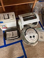 Four kitchen appliances on carpet: Toastmaster bread maker with manual, Kenmore Choice Jumbo Electric Frying Pan with manual, George Foreman rotisserie cooker, glass Savoureux convection oven.