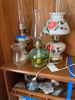 Photo showing two oil lamps, one electric lamp with floral design, and some decorative rocks on wooden shelf.