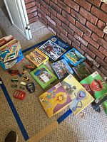 Photo showing assorted toys including Fisher-Price play desk box, several jigsaw puzzles, and small toy cars on carpeted floor against a brick wall