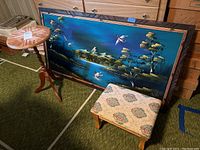 Overview of lot items including a retro velvet painting, upholstered stool, wood side table, and plant stand on green carpet.