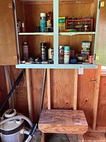Wooden wall cabinet containing assorted spray paints, cleaners, lubricants on shelves above wooden bench with paint marks, vacuum hose visible.