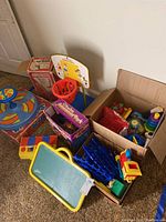Lot of various vintage toddler toys including Sit 'n' Spin, chalkboard, basketball goal, and toy train with tracks arranged on floor next to wall