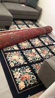 Full view of rectangular wool area rug rolled partially, showing floral pattern with dark background, placed on floor near furniture.