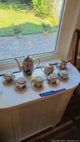 Full set displayed on white surface by window including teapot, sugar bowl, creamer, 5 cups and 5 saucers setup in a neat arrangement