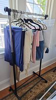 Photo shows metal clothing rack with rolling wheels and adjustable height. Several garments hang on the chrome bar but are not included.