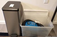 Photo showing the gray Curver step trash bin with black foot pedal beside a clear plastic tote bin containing a blue Eureka Rally vacuum.