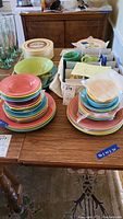 Stack of colorful Fiesta plates, bowls, and cups arranged on a wooden table, showing various colors and sizes.